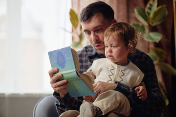 Caucasian man reading Hanukkah storybook to toddler child while sitting together, both focused on...