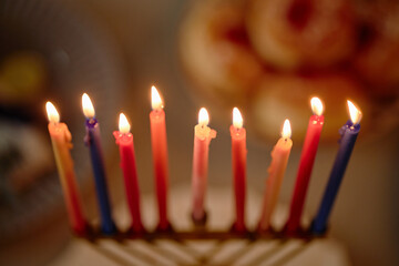 Menorah with nine colorful candles burning, celebrating Hanukkah tradition, close up of flames and wax melting, blurred background with festive food visible in soft focus