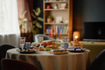 Hanukkah table setting featuring menorah with colorful candles, traditional foods including sufganiyot and latkes, glassware and plates arranged for festive Jewish holiday meal