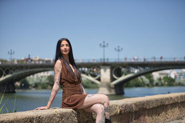 Young woman, brunette, tall, thin and very tattooed, with brown dress, looking to infinity sitting by the river with the Triana bridge in the background. Concept of youth, millennial.