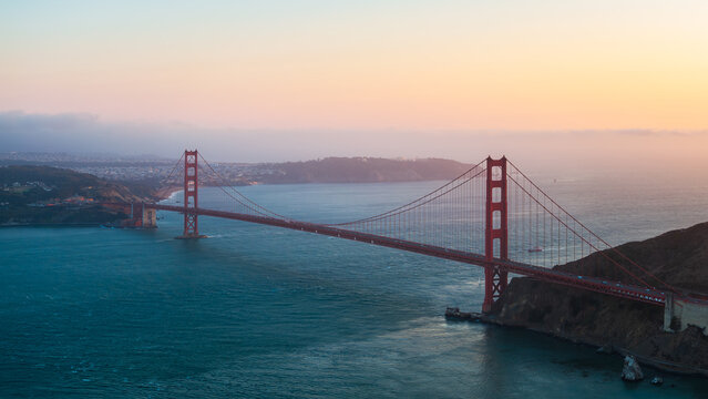 Aerial view of the iconic Golden Gate Bridge painted in warm sunset hues, standing tall above the tranquil, deep blue waters of the bay, San Francisco, California, United States.