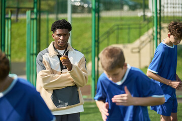 Black man standing on sports field holding stopwatch observing group of teenage boys running in blue uniforms during outdoor athletic training session