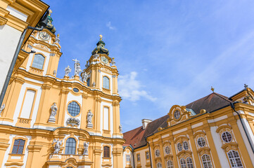 Melk Abbey showcases impressive Baroque architecture with golden accents against a bright blue sky, featuring ornate detailing and towering spires immersed in sunlight, complemented by the