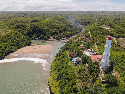 Aerial view of a white lighthouse standing tall near a river mouth, surrounded by lush green forest, Java, Indonesia.