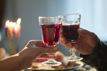 Adult hands holding wine glasses and making toast near lit Hanukkah menorah, celebrating Jewish holiday with traditional festive meal in background