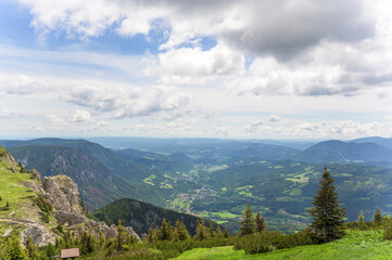 Fototapeta premium The image showcases a breathtaking panoramic view from Rax in Austria featuring lush green valleys and towering mountains, with steep, rocky outcrops in the foreground and a serene landscape