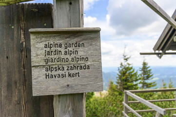 A weathered wooden sign at Rax Austria displays text against a backdrop of green trees and distant hills under a clear sky with soft clouds, showcasing the serene mood