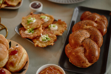 Traditional Hanukkah foods including braided challah bread, potato latkes topped with sour cream and green onions, jelly filled donuts and assorted fruit preserves arranged on table