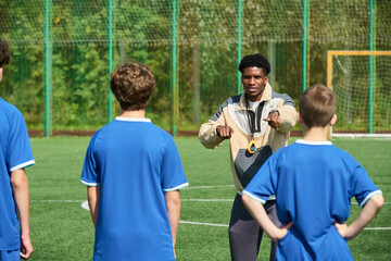 Black man instructing group of teenage boys standing on soccer field during outdoor sports training session, coach holding whistle and gesturing toward players