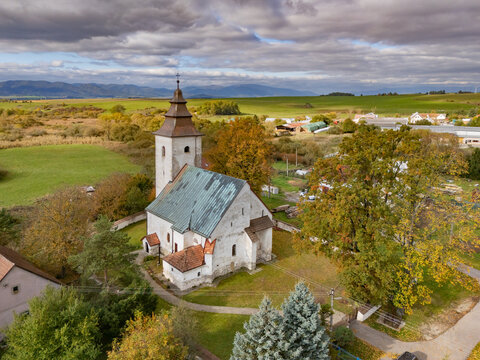 Aerial view of a medieval church with a tall tower nestled among trees displaying vibrant autumn foliage, contrasting with the open fields beyond, H&Atilde;&iexcl;j, &Aring;&frac12;ilina Region, Slovakia.