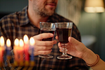Middle aged Caucasian man and woman clinking wine glasses during Hanukkah celebration, holding drinks near lit menorah candles, hands and faces partially visible, festive setting