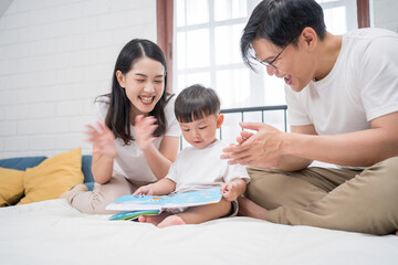 Asian family reading picture book together on bed in cozy bedroom. Parents guiding toddler boy with smiles. Concept of early childhood education, bonding, learning at home, and family love.