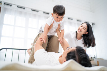 Fototapeta premium Asian family spending quality time together at home, father helping toddler boy dress on bed while mother watches with smile. Concept of parenthood, care, and morning family routine in cozy home.