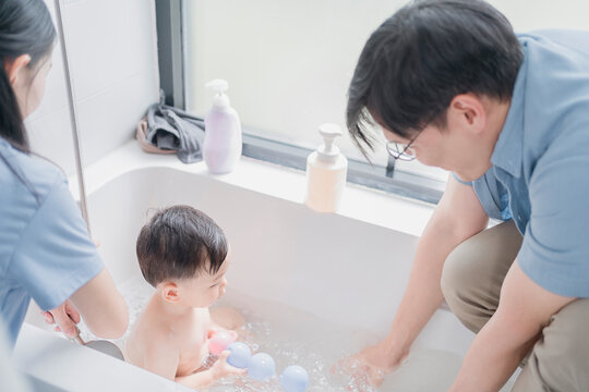 Asian parents bathing toddler boy in a bathtub, playing with colorful balls. Family hygiene care, bonding time, and daily parenting routine in bright, clean bathroom environment. - Powered by Adobe