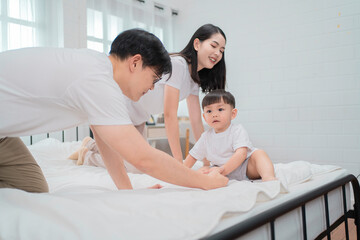 Fototapeta premium Happy Asian family enjoying time together in cozy bedroom. Parents playing with their toddler son on bed, smiling and bonding. Concept of love, connection, and joyful parenting at home.