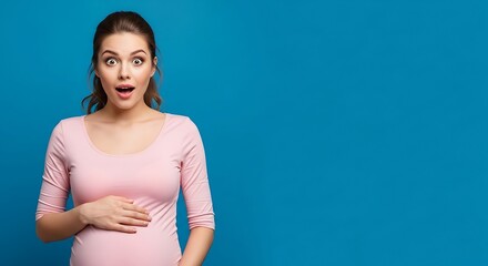 A lovely pregnant woman expresses genuine surprise and excitement while gently cradling her growing baby bump against a vibrant blue background.