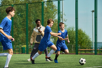 Obraz premium Group of teenage boys running and competing for soccer ball on outdoor field while Black male coach observing and instructing during training session