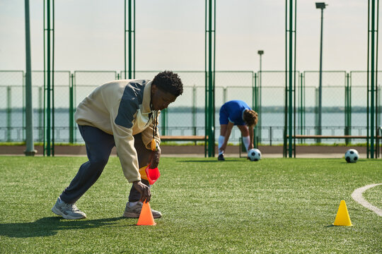 Black man arranging orange cones on outdoor soccer field while teenager in blue sportswear stretching in background, soccer balls scattered on artificial turf - Powered by Adobe