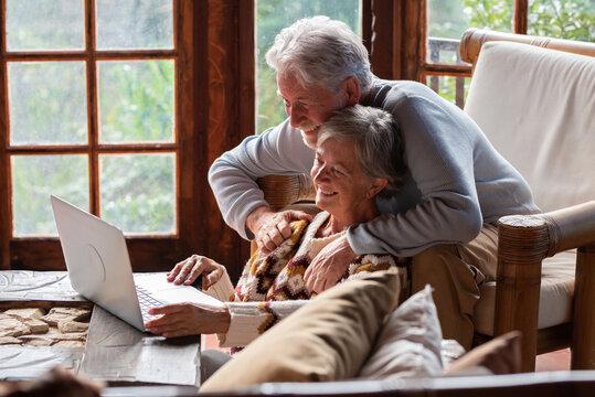 Smiling elderly couple using laptop together at home in a warm rustic chalet-style interior, enjoying leisure time, remote work, or video call, concept of digital lifestyle, togetherness, and happines