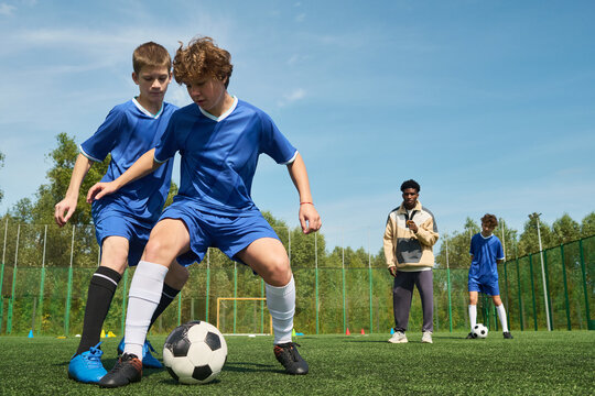 Two teenage boys competing for soccer ball on outdoor field while Black male coach observing in background, another teenage boy standing with soccer ball - Powered by Adobe