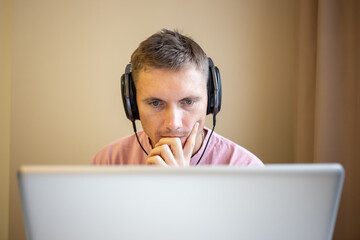 A freelance guy in a pink T-shirt is working on a laptop, wearing headphones, doing remote work from home. A man uses a laptop to work remotely at home while listening to music with headphones. 