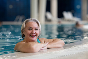 Smiling senior woman enjoying relaxation in an indoor swimming pool, leaning on the poolside. Concept of active lifestyle, wellness, health, hydrotherapy, and leisure for older adults.