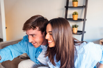 Modern couple sitting on the sofa at home using a laptop together, smiling and laughing during...