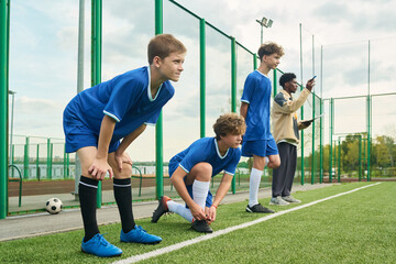 Three teenage boys preparing for soccer training on outdoor field, standing and crouching near sideline while Black man coach giving instructions in background