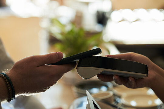 Caucasian young adult man holding smartphone making contactless payment using wireless terminal held by another , closeup of hands performing digital transaction in cafe