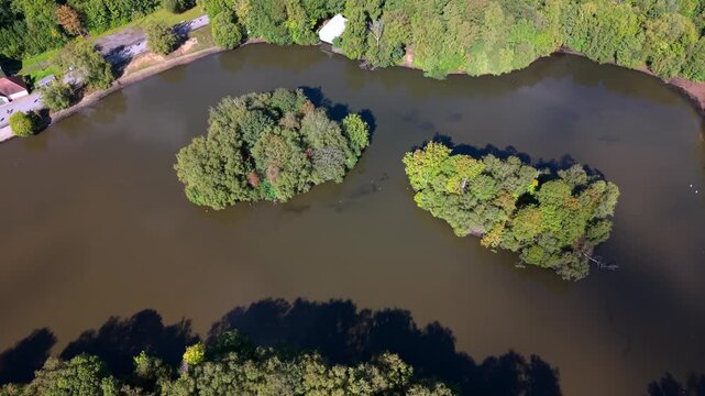 Bird&rsquo;s-eye aerial video of Heaton Park boating lake with small wooded islands surrounded by trees. Prestwich, Manchester. 