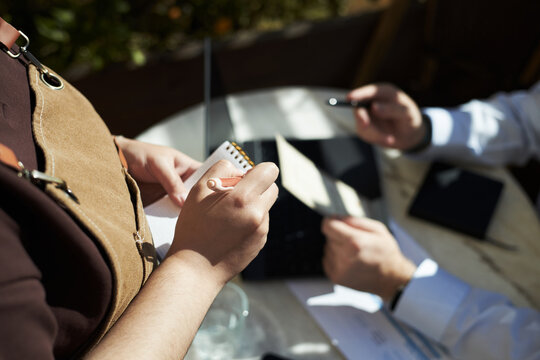 Caucasian young adult woman holding notepad taking order from Caucasian middle aged man sitting at table using laptop and holding pen, outdoor cafe setting, hands visible