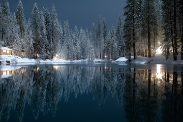 Serene winter landscape featuring a tranquil lake reflecting snow-covered trees and a cozy cabin illuminated by soft lights, creating a peaceful nighttime atmosphere