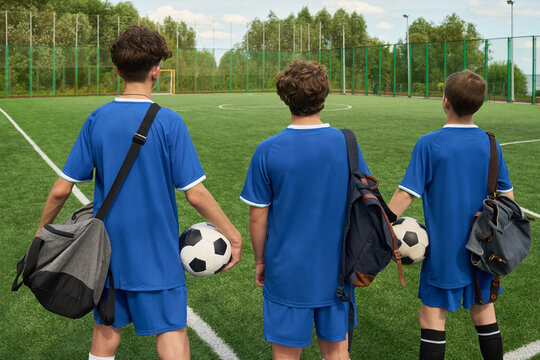 Three teenage boys standing on soccer field holding soccer balls and sports bags, facing away from camera, preparing for soccer practice or match outdoors - Powered by Adobe
