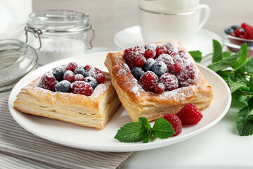 Tasty puff pastries with berries, powdered sugar and mint on white table, closeup