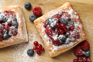 Tasty puff pastries with berries and powdered sugar on wooden table, flat lay