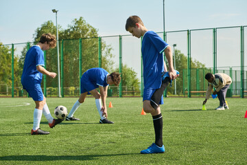 Group of and Black teenagers warming up and stretching on outdoor soccer field before practice, one teenager balancing soccer ball on foot, green fence and trees in background