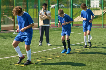Three teenage boys performing agility ladder drills on outdoor sports field while Black man coach observing and instructing in background during soccer training session