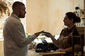 Caucasian young adult man making contactless payment with smartphone while Hispanic young adult woman holding card reader at counter in cafe setting, both focused on transaction