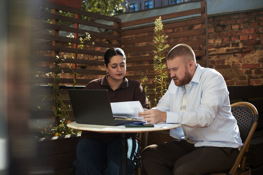 Caucasian middle aged man and Caucasian young adult woman sitting at outdoor table discussing documents with laptop open, both focused on paperwork during business meeting