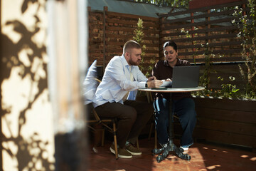 Caucasian young adult man and Middle Eastern young adult woman sitting at outdoor table working together on laptop, both focused on screen, discussing project in garden setting