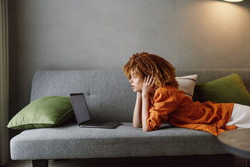 Young woman with curly hair relaxes on a sofa while using a laptop, dressed in an orange top, in a cozy setting with green cushions, reflecting modern lifestyle