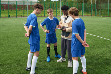Obraz premium Teenage boys standing on soccer field listening to Black male coach explaining strategy using digital tablet during outdoor team practice session