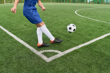 Teenager boy with appearance kicking soccer ball near corner line on outdoor football field, lower body visible, wearing sports uniform and cleats during game