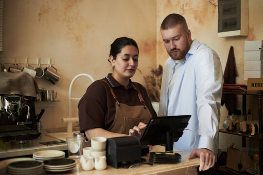 Caucasian young adult man and Hispanic young adult woman standing at counter using touchscreen point of sale terminal in coffee shop, both focusing on digital transaction process