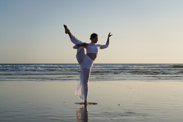 Model in white clothing performing a high dance or yoga pose on a beach near the ocean. Shot at sunrise or sunset. Female dancer on beach.