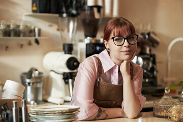 Portrait of young adult Caucasian woman with glasses and tattooed arm leaning on counter in coffee shop, resting chin on hand and looking into camera with relaxed expression