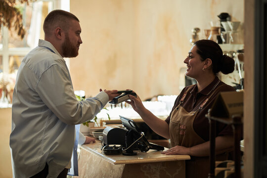 Caucasian young adult man paying with smartphone while smiling Middle Eastern young adult woman barista processing transaction at counter in modern coffee shop - Powered by Adobe