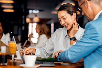Asian woman and coworker talking about work while using laptop in modern coffee shop.