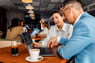 Asian woman and coworker talking about work while using laptop in modern coffee shop.