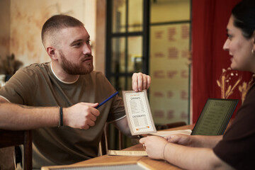 Caucasian young adult man discussing menu options with Caucasian young adult woman in restaurant, man holding menu and gesturing with pen while woman listening attentively